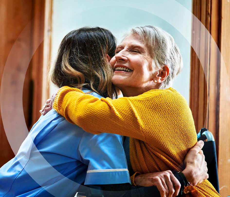Young woman in scrubs hugging an older woman in a wheelchair, conveying care and support.