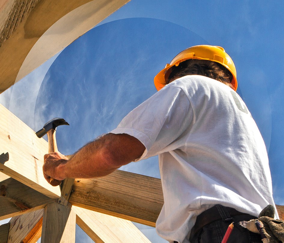Man in a yellow hard hat hammering a nail while working on a house, representing housing construction.